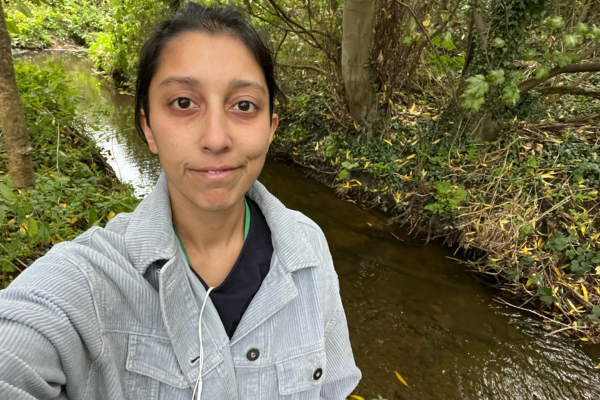 Selfie of a dark-hair woman wearing a grey jacket standing in front of a river background